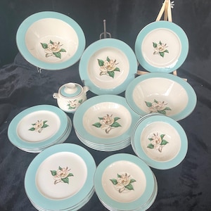 May include: A vintage dinnerware set featuring a white magnolia flower design on a cream background with a light blue rim. The set includes plates, bowls, and a creamer.