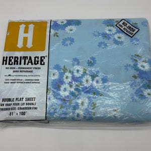 May include: A Heritage brand double flat sheet in original plastic packaging. The fabric is light blue with a white and dark blue floral daisy pattern. The label indicates 50% polyester and 50% cotton, with a finished size of 205.7 cm by 254 cm.