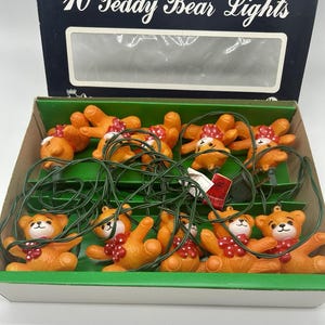 May include: A set of 10 teddy bear lights in a cardboard box labeled 10 Teddy Bear Lights. Each light features a small orange plastic bear with a white face and a red polka dot bow tie, connected by a green electrical cord with a plug.