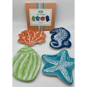 May include: A set of four ceramic plates shaped like marine life. The plates are in the shapes of a seahorse, starfish, shell, and coral. The plates are in shades of blue, green, orange, and white. The box reads "Sea Breeze plate set of four."