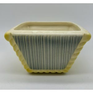 May include: A square ceramic planter with a ribbed, pale blue exterior and a cream-coloured interior. The edges are trimmed with a yellow, textured design. Suitable for small plants or succulents.
