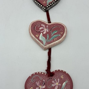 May include: A vertical hanging decoration consisting of three heart-shaped wooden plaques connected by a dark red fabric ribbon. Each heart is painted a dusty rose color with hand-painted light green leaves, white flowers, and small yellow dots.