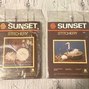 Peut inclure: Deux kits Sunset Stitchery dans un emballage plastique. L'un présente un motif floral, l'autre une oie dans un nid. Les deux kits ont une marque marron et orange avec le mot "Sunset" en haut.