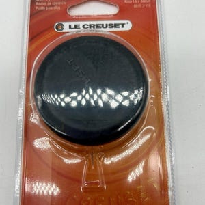 May include: A black Le Creuset replacement cookware knob packaged in a clear plastic blister pack with an orange cardboard backing. The packaging features the Le Creuset logo and text in multiple languages, including Cookware Knob and Perilla para ollas.