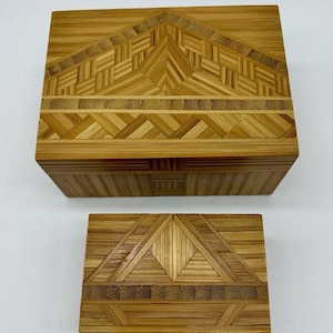 May include: Two rectangular wooden boxes featuring a light brown natural wood grain and intricate geometric straw marquetry patterns on the lids. The larger box is positioned above the smaller box, both displaying detailed woven straw designs.