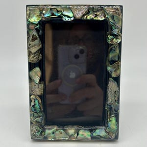 May include: Rectangular black photo frame with iridescent abalone shell accents. The frame has a glossy finish and a dark interior, suitable for displaying a photograph. The abalone shell pieces are in shades of green, blue, and silver.