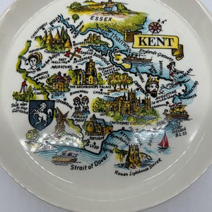 May include: A colorful ceramic plate depicting a map of Kent, England. The plate features various landmarks and points of interest, including Canterbury Cathedral, Dover Castle, and the River Thames. The plate is decorated with a blue and white border.