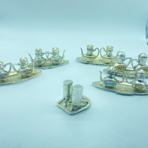 May include: Set of silver salt and pepper shakers in the shape of teapots. Each shaker has a different city name on it, including Dauphin Man, Brandon, Winnipeg, and Vancouver BC. The shakers are on small silver trays.