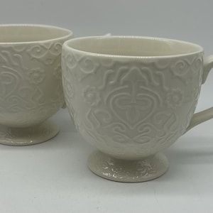 May include: Two off-white ceramic mugs with embossed floral and scrollwork patterns. Each mug has a pedestal base and a curved handle. The mugs are displayed on a white surface.