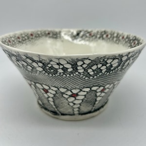May include: A ceramic bowl featuring a black and white lace-like pattern across the exterior. The design includes intricate floral motifs with small, distinct red accents centered within the petals, set against a smooth, off-white background.