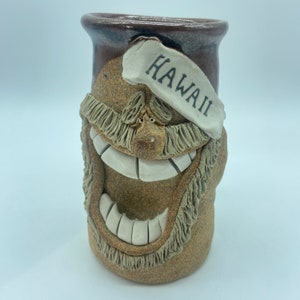 Peut inclure: Une tasse en céramique brune avec un visage souriant et une bannière blanche qui dit "Hawaii". La tasse a un glaçage brun et une barbe blanche.