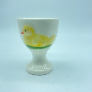 May include: A white ceramic egg cup featuring a yellow duck design. The duck has a red beak and black eyes.