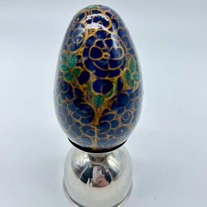May include: A blue and gold decorative egg with a floral pattern sits on a silver stand. The egg is hand-painted with intricate details.