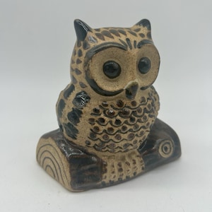 May include: A ceramic owl figurine perched on a horizontal log. The owl is tan with dark brown painted details on its feathers, large black eyes, and a small beak. The log base is dark brown with lighter tan circular rings on the side.