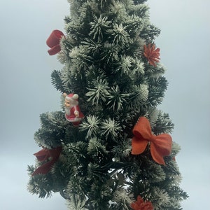 May include: A small artificial Christmas tree with a flocked finish, decorated with red bows, red and white Santa figurines, and orange felt flowers. The tree is on a black metal stand.
