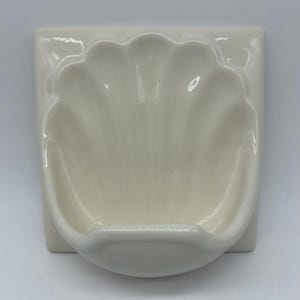 May include: A glossy off-white ceramic wall-mounted soap dish featuring a molded shell design. The piece has a square backplate and a curved, scalloped basin that extends outward, providing a functional holder for a bar of soap.