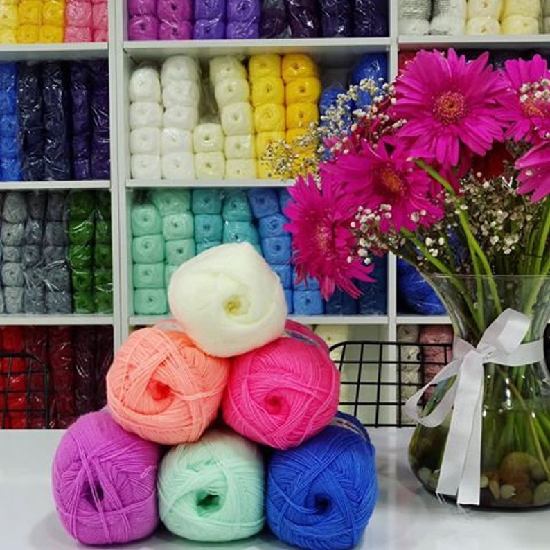 May include: A collection of colorful yarn balls in various shades of pink, blue, green, white, and orange. The yarn is arranged in a semi-circle on a table with a bouquet of pink flowers in a vase in the background.
