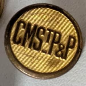 May include: A circular gold-colored button featuring a textured, horizontally lined surface. The letters CMSTP&P are embossed in a bold, dark font across the center of the button, which has a raised, smooth, and rounded outer edge.