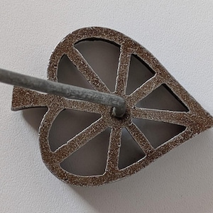 May include: A leaf-shaped, metal rosette iron with a handle. The iron has a detailed, openwork design with a textured, brown finish. The handle is a simple, gray metal rod.
