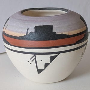 May include: A small, round ceramic vase with a wide opening. The vase features a Southwestern design with a white base, a black and red band, and a painted landscape scene in shades of gray, orange, and black. The vase is approximately 4 inches in diameter.