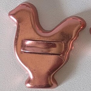 May include: A metallic copper-colored chicken-shaped object with a smooth, reflective surface. The figure features a distinct head, a rounded body, and a small base. A horizontal rectangular bar is embossed across the center of the chicken's body.