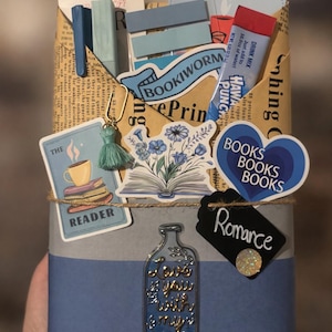 May include: A rectangular package wrapped in newspaper print paper, secured with twine. It features stickers of a coffee cup, blue flowers, and a heart reading BOOKS BOOKS BOOKS. A bottle-shaped charm reads Love you with my whole, with a Romance tag.