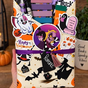 May include: A rectangular gift package wrapped in cream paper with Halloween motifs, featuring a purple ribbon, a black tag labeled Romance with a tassel, and various stickers including a ghost with a scythe, a cartoon couple, and a tombstone.