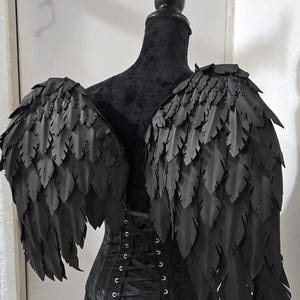 Puede incluir: Un par de alas grandes de color negro con capas estilo pluma, unidas a un corsé negro con cierre de cordones en la espalda. Las alas presentan varias filas de segmentos negros puntiagudos y superpuestos, y el corsé tiene un acabado de tela oscura.