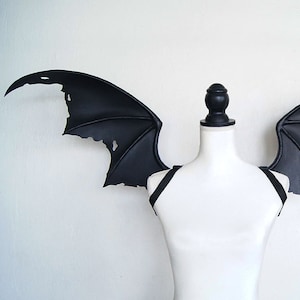 May include: Black leather bat wings with a distressed look, attached to a black harness. The wings are displayed on a white mannequin.