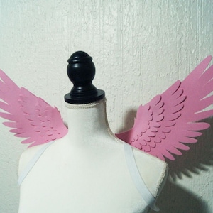 Puede incluir: Alas de ángel de papel rosa unidas a un torso de maniquí blanco. Las alas están detalladas con un borde festoneado y un diseño de plumas.