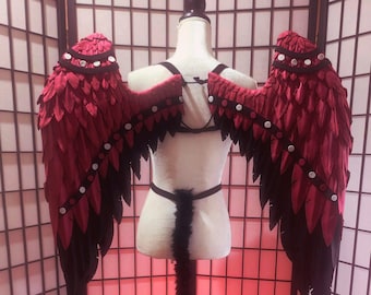 May include: Red and black costume wings with white accents, displayed on a mannequin. The wings have a layered feather design and are attached to a harness. A matching tail is also visible, creating a complete costume.