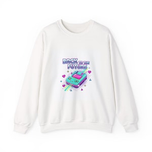 Puede incluir: Sudadera blanca con un gráfico de consola de videojuegos retro y el texto "BACK TO THE FUTURE". El diseño incluye una consola turquesa y morada con acentos coloridos, corazones y formas geométricas.