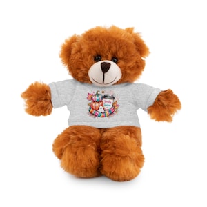Puede incluir: Un oso de peluche marrón que lleva una camiseta gris claro. La camiseta presenta un gráfico colorido de calabazas apiladas, una taza de café con el texto Spooky Season, un pequeño fantasma blanco y una bufanda a rayas rodeada de hojas de otoño.