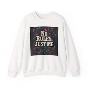 Puede incluir: Sudadera blanca con un diseño gráfico cuadrado negro. El diseño presenta el texto "NO RULES, JUST ME" en una fuente lúdica, rodeado de ilustraciones rosas y azules de imperdibles, corazones y cuadrados. Una prenda casual y cómoda.