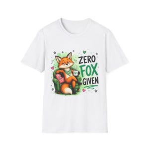 Puede incluir: Una camiseta blanca de manga corta con un gráfico de un zorro naranja sentado en un sillón verde, sosteniendo una taza de café. El zorro lleva un pañuelo verde y está sentado junto a un cojín rosa con la etiqueta NOT TODAY. El texto ZERO FOX GIVEN está impreso en negro y verde.