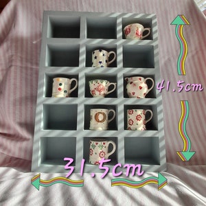 May include: A gray wooden shelf with 12 square compartments, each holding a small ceramic mug or pitcher. The mugs and pitchers feature various colorful patterns and designs. The shelf measures 31.5 inches wide and 41.5 inches tall.