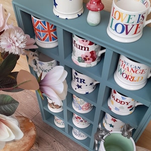 Könnte beinhalten: Ein blaues Holzregal mit 12 Fächern, in denen jeweils eine weiße Keramiktasse mit einem anderen Design steht. Die Tassen zeigen verschiedene bunte Designs, darunter Sterne, Blumen, Herzen und Wörter wie "Love is Love", "Mum", "Mr", "Daddy" und "We Can Change the World".