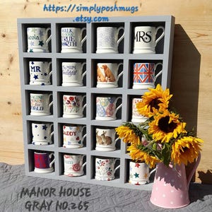 Puede incluir: Una estantería de madera gris con 18 compartimentos cuadrados, cada uno con una taza de diferentes patrones. La estantería está etiquetada como "MANOR HOUSE GRAY NO. 265". Una regadera rosa con lunares blancos está en primer plano.