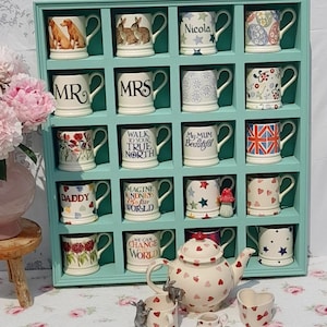 Puede incluir: Una colección de tazas de cerámica, una tetera y una jarrita de crema exhibidas en una estantería de madera de color turquesa. Las tazas presentan varios diseños, incluyendo patrones florales, texto como "MR" y "MRS", y una bandera británica. La tetera y la jarrita de crema tienen un patrón de corazón.