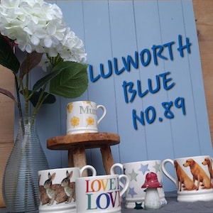 Puede incluir: Una colección de tazas de cerámica con varios diseños, incluyendo conejos, perros, estrellas y motivos florales. Una taza tiene la palabra "Mum" y flores amarillas. El fondo presenta un letrero azul con el texto "LULWORTH BLUE NO. 89". También hay un jarrón con flores blancas.
