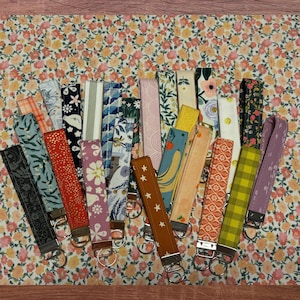 May include: A collection of 20 colorful fabric keychains with various patterns and designs. The keychains are attached to silver key rings.