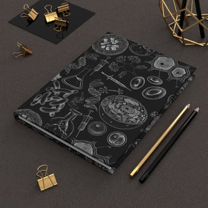 以下が含まれることがあります： 細胞、DNA、実験器具を特徴とする白い科学イラストパターンが施された黒いジャーナル。金と黒の鉛筆と金のペーパークリップが、濃い灰色の表面にあるジャーナルの隣にあります。