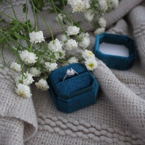 May include: A teal velvet ring box with a silver ring with a clear stone in the center. The box is sitting on a light gray knitted fabric with white flowers.