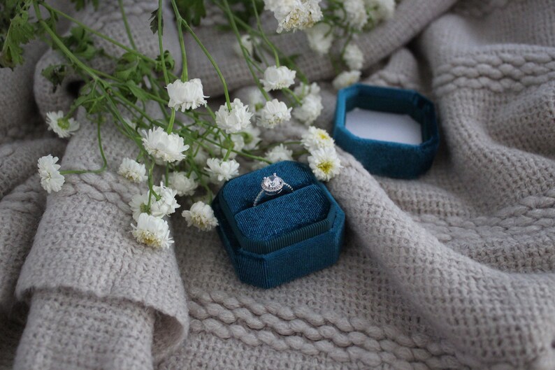 May include: A teal velvet ring box with a silver ring with a clear stone in the center. The box is sitting on a light gray knitted fabric with white flowers.