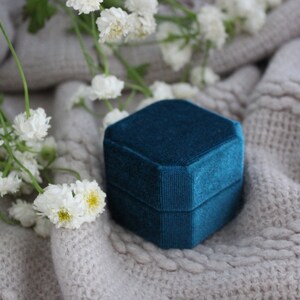 May include: A teal blue velvet ring box on a white knitted surface with white flowers.