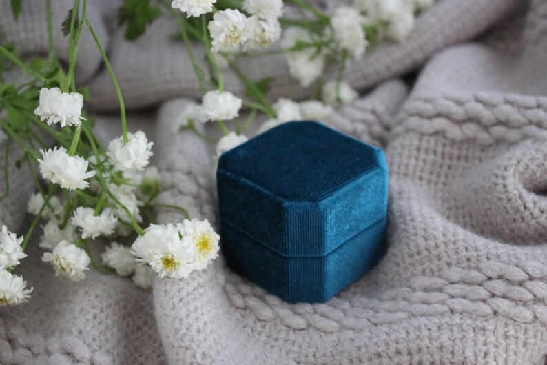 May include: A teal blue velvet ring box on a white knitted surface with white flowers.