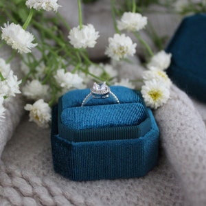 May include: A teal velvet ring box with a silver ring with a clear stone in the center. The box is sitting on a white fuzzy surface.