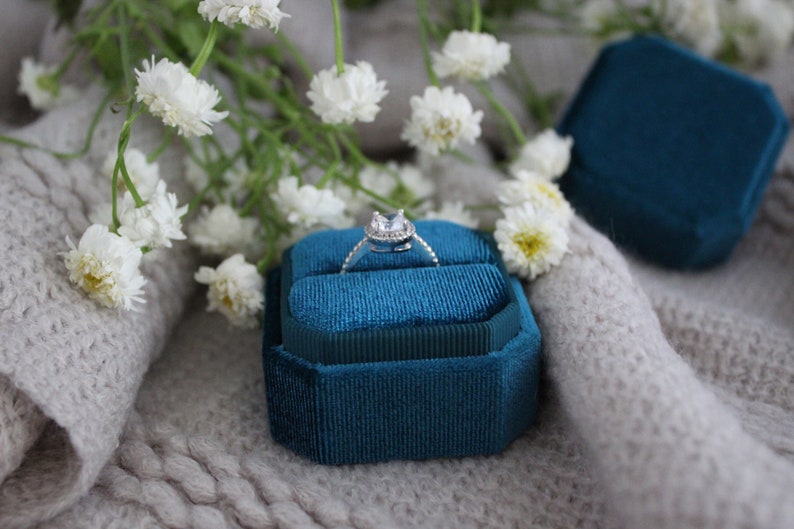 May include: A teal velvet ring box with a silver ring with a clear stone in the center. The box is sitting on a white fuzzy surface.