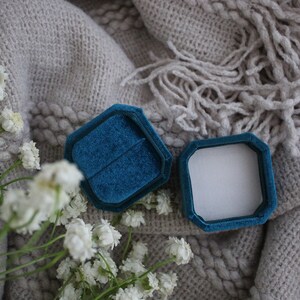 May include: A teal blue velvet ring box with a white interior, open and lying on a gray knitted blanket with white flowers.