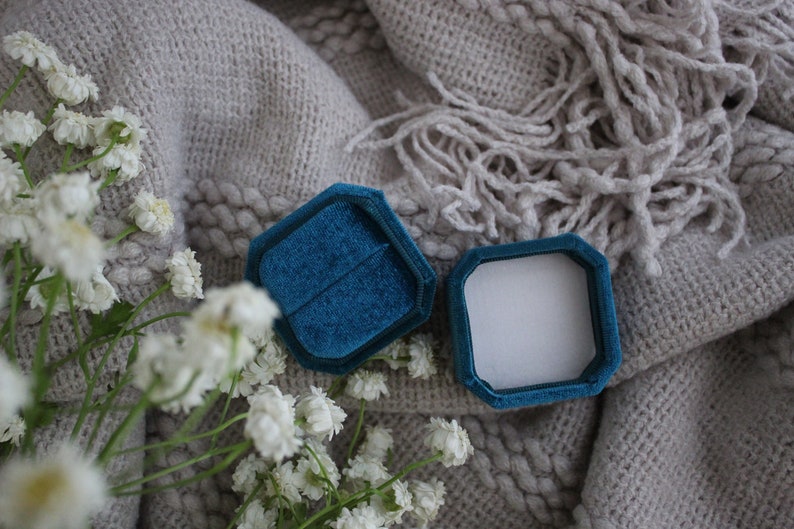 May include: A teal blue velvet ring box with a white interior, open and lying on a gray knitted blanket with white flowers.